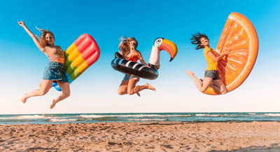 Three women with pool inflatables jumping in the air at the beach.